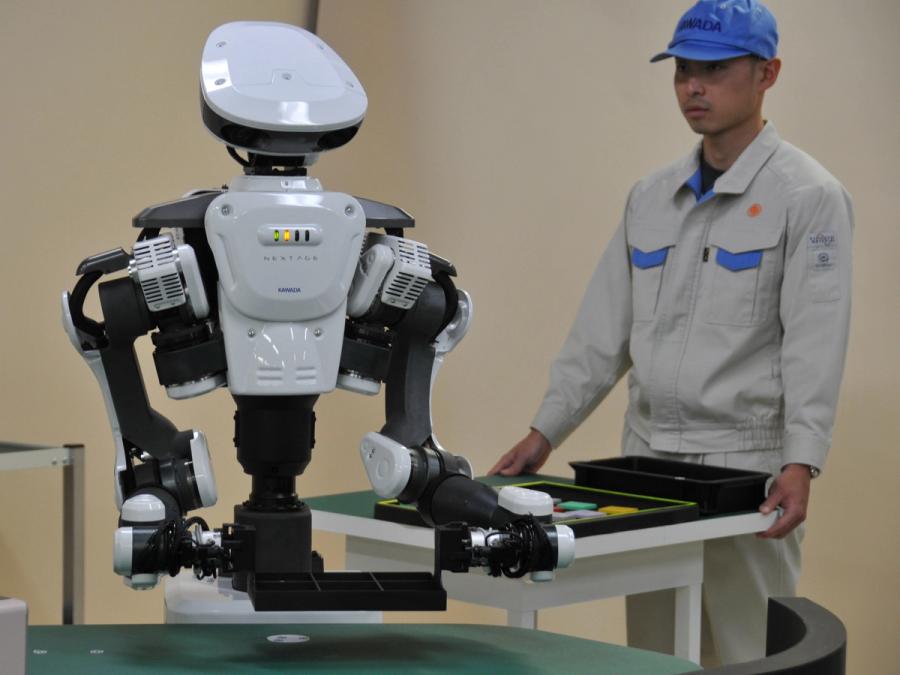 The robot and a human clothed in a hat and worker jumpsuit stand in front of a cart.