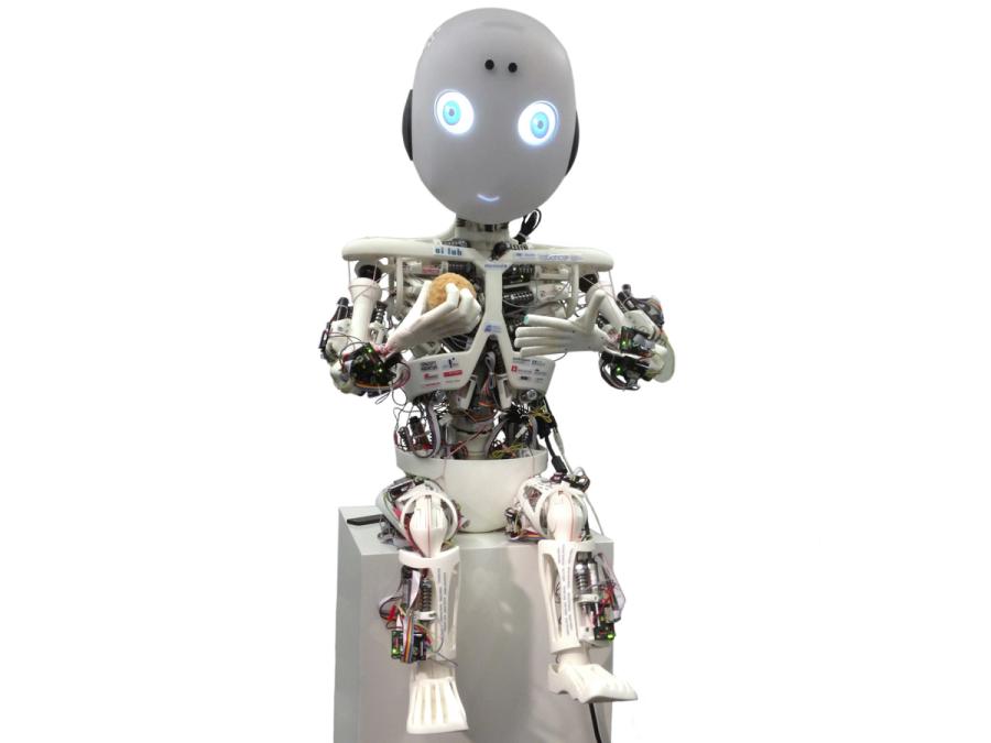 A child-like skeletal robot sits on a block. It's musculature and electronics are exposed. Its head is egg shaped with glowing blue eyes and mouth.