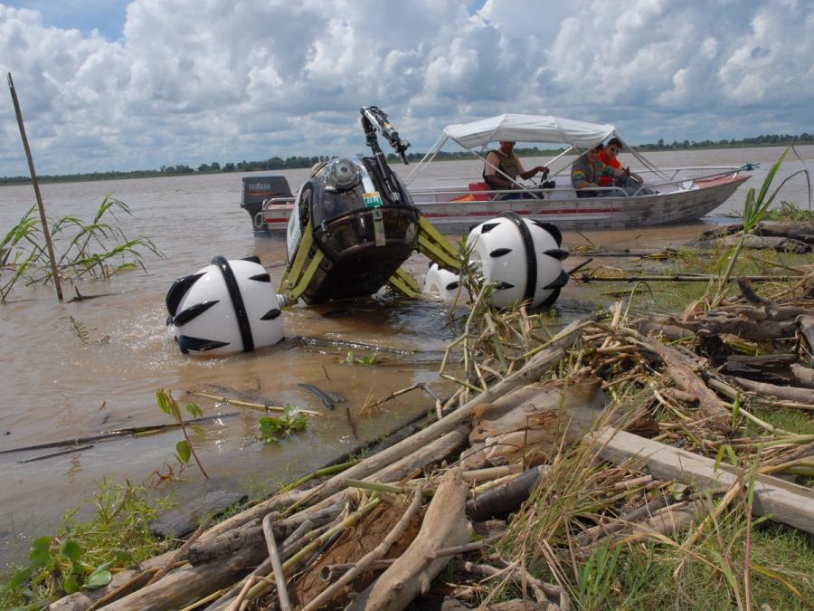 The robotic vehicle is in the water next to a boat and near shore covered in wood.