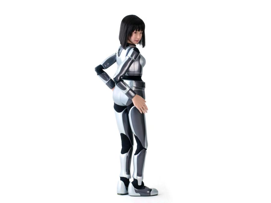 Full body rear view of the robot. Its body is encased in a silver and black covering, but it's hands and face have the appearance of flesh. It poses with hands on its hips.