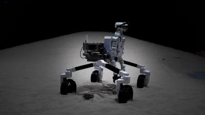 Space robot GITAI G1, a humanoid with a white metal body with a sensor-packed head and articulated arms, mounted on a rover-like wheel base, drives over a sandy simulated lunar environment.