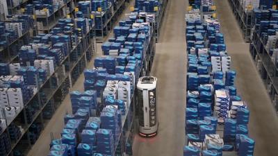 A warehouse seen from above with multiple tall racks of shoe boxes, with a mobile robot the size of a refrigerator driving down a row of racks.