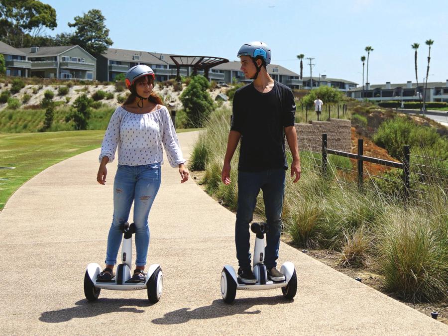 Two people in helmets ride mini Segways which include the base on two wheels and a vertical piece balanced between the rider's legs.