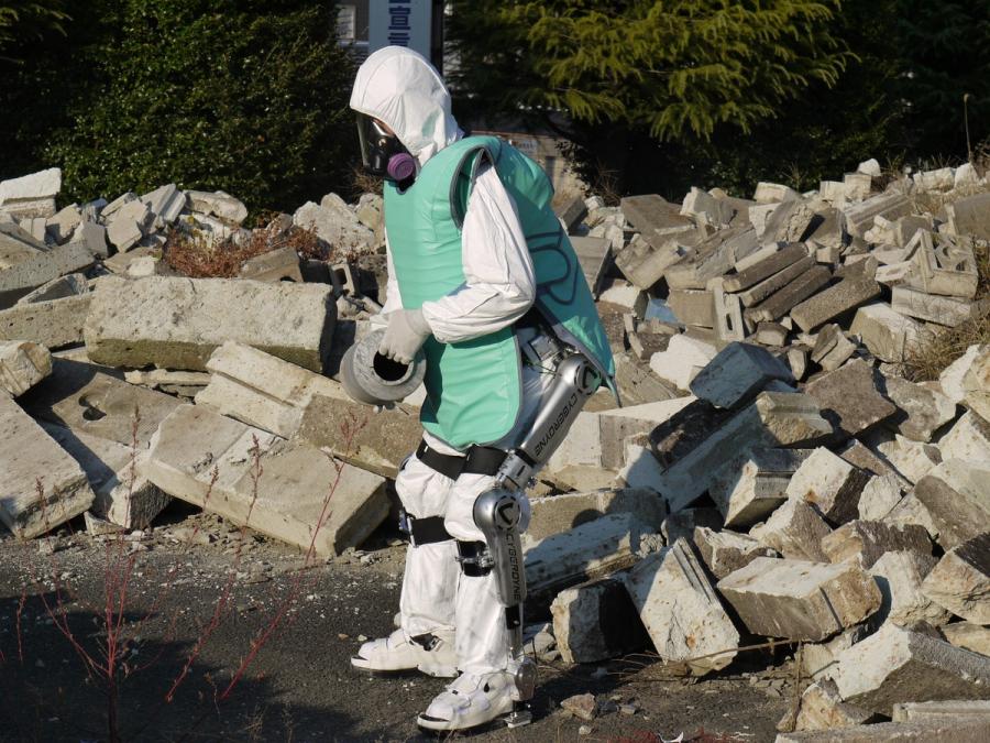 A person in a clean room bunny suit, gloves, dust mask, x-ray shield, lower-body exoskeleton suit stands outside amid rubble.