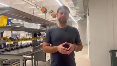 A man with dark hair, short beard, wearing a gray tshirt looks at the camera while standing in an electronics lab with benches and components in containers.